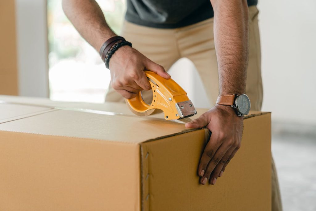 pexels-photo-4246120 Crop faceless young male with wristwatch using adhesive tape while preparing cardboard box for transportation